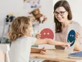Dziecko i pedagog pracują nad emocjami podczas terapii pedagogicznej, używając kart z buźkami.