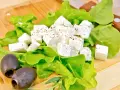 Kostki sera feta, przyprawione ziołami, leżą na liściach sałaty obok czarnych oliwek. Idealne na lekki posiłek.
