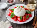 Burrata z pomidorami i bazylią na talerzu, obok pieczywo i przyprawy.
