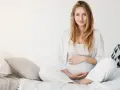 Uśmiechnięta kobieta w ciąży w białych ubraniach siedzi na łóżku, delikatnie trzymając się za brzuch.