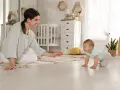 Uśmiechnięta mama zachęca do pierwszych kroków, kiedy dziecko raczkuje po dywanie w pokoju dziecięcym.
