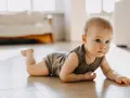 Dziecko w body leży na brzuchu na drewnianej podłodze, patrząc w bok.