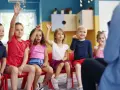 Dzieci na czerwonych krzesłach podnoszą ręce, jakby odpowiadały na pytanie, czy zerówka jest obowiązkowa.