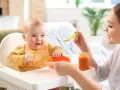 Uśmiechnięte 2 miesięczne dziecko w żółtym ubranku w kropki siedzi w krzesełku do karmienia. Mama podaje mu jedzenie z łyżeczki.