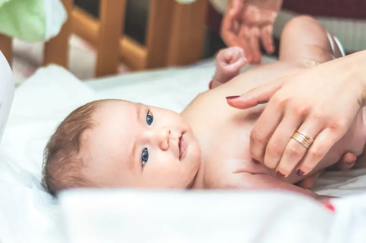Niebieskooki bobas leży na białym prześcieradle, a jego mama delikatnie go dotyka. Na palcu mamy widać obrączkę, a na jej paznokciach ciemny lakier. Dziecko ma na sobie tylko pieluszkę, a jego skóra jest gładka i delikatna, jak **perły Epsteina**.