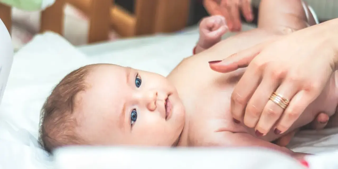 Niebieskooki bobas leży na białym prześcieradle, a jego mama delikatnie go dotyka. Na palcu mamy widać obrączkę, a na jej paznokciach ciemny lakier. Dziecko ma na sobie tylko pieluszkę, a jego skóra jest gładka i delikatna, jak **perły Epsteina**.