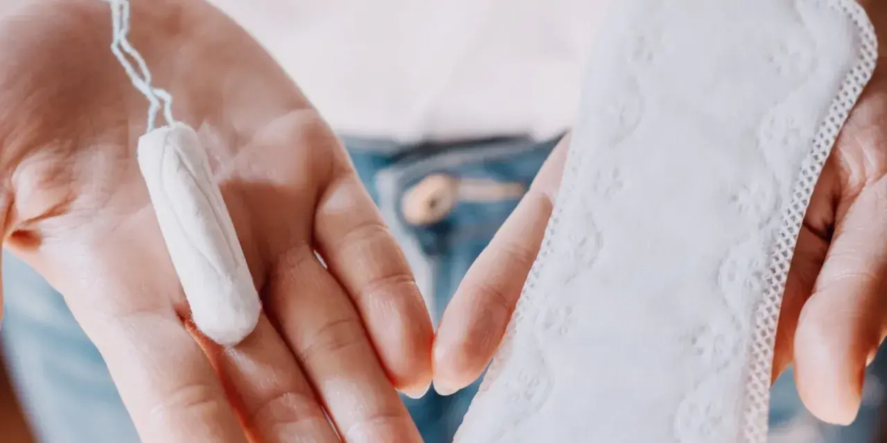 Dłonie trzymają tampon i podpaskę. Czasem pojawia się plamienie zamiast okresu, wtedy wybór jest ważny.