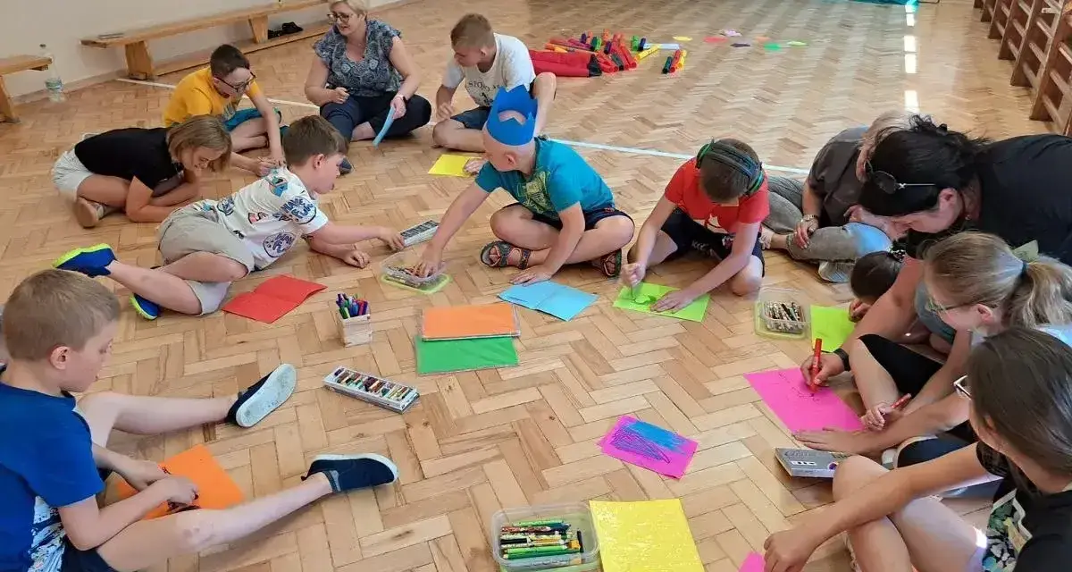 Dzieci i dorośli siedzą na podłodze, rysując i bawiąc się. Wszyscy są zaangażowani w zabawy integracyjne, tworząc kolorowe prace.