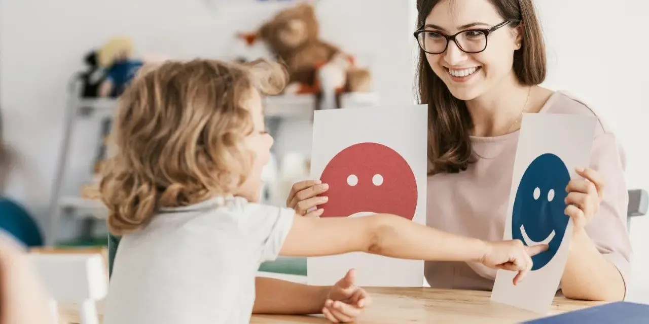 Dziecko i pedagog pracują nad emocjami podczas terapii pedagogicznej, używając kart z buźkami.
