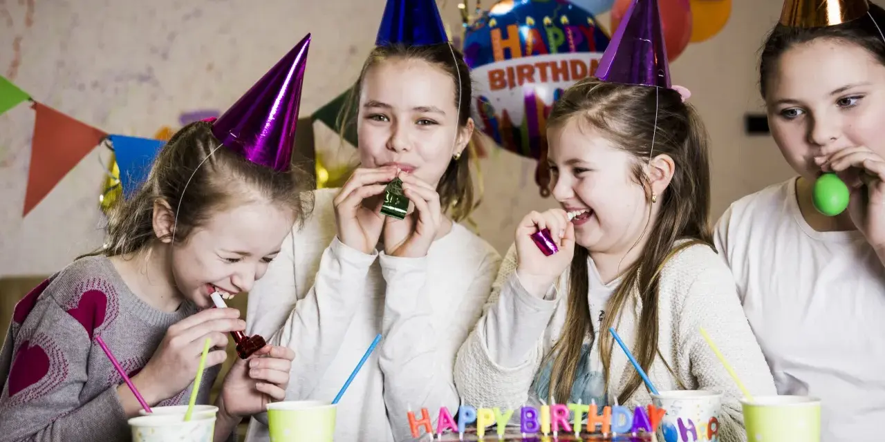 Radosne zabawy na urodziny! Dziewczynki w stożkowych czapkach dmuchają w gwizdki, śmiejąc się przy torcie z napisem "Happy Birthday".