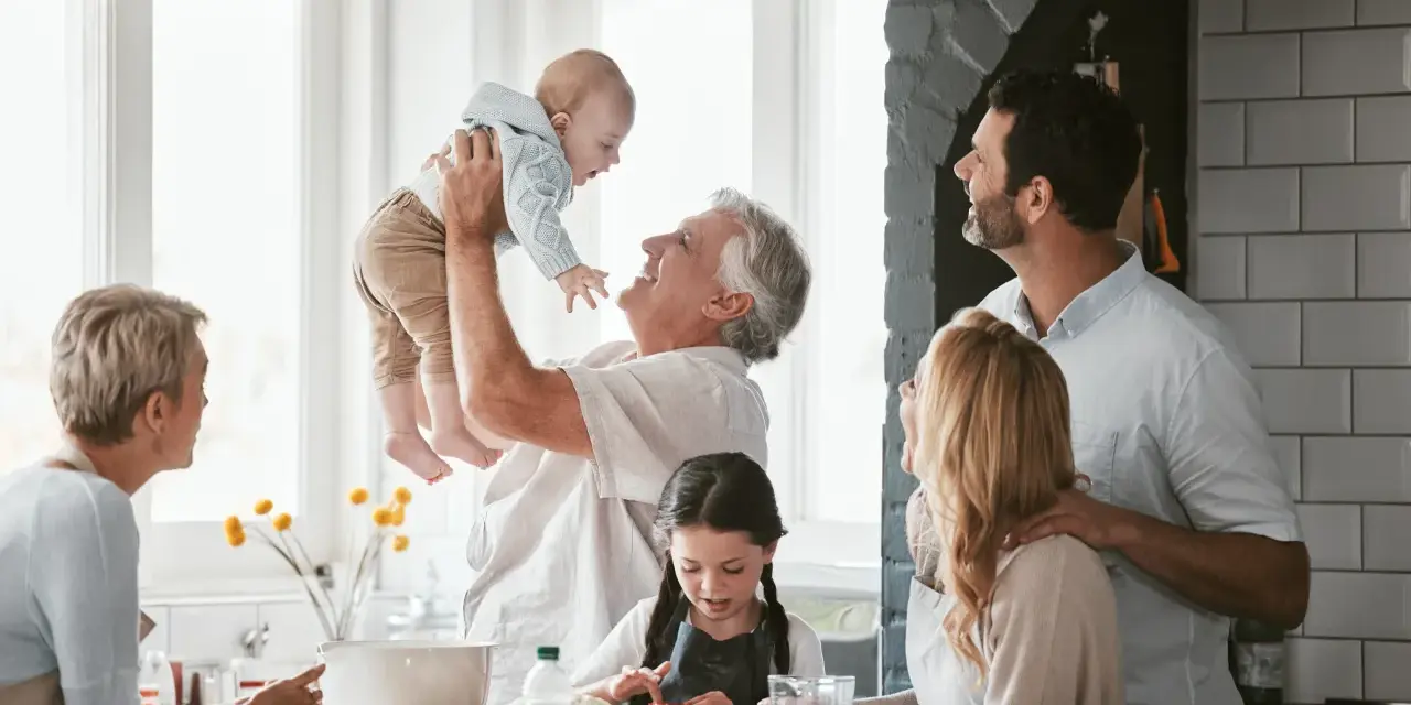 Rodzina w kuchni. Dziadek podrzuca wnuka, babcia patrzy z uśmiechem. Dzieci pomagają w przygotowaniach. To są babciowe dla kogo? Dla wszystkich, którzy kochają wspólne chwile.
