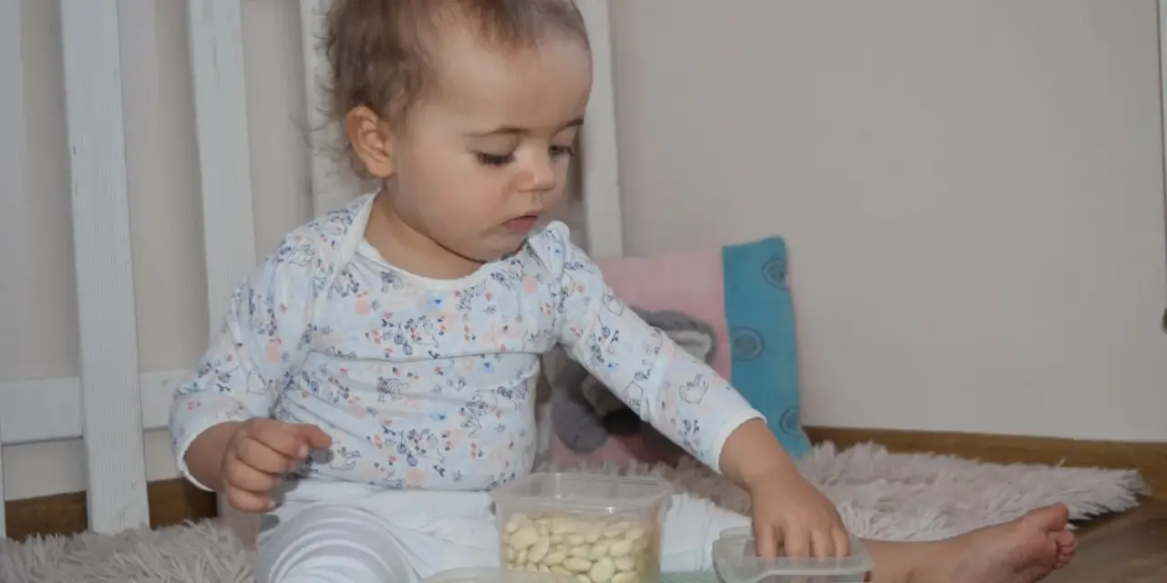 Roczne dziecko bawi się fasolkami w pojemnikach. Proste zabawy dla rocznego dziecka rozwijają motorykę małą.