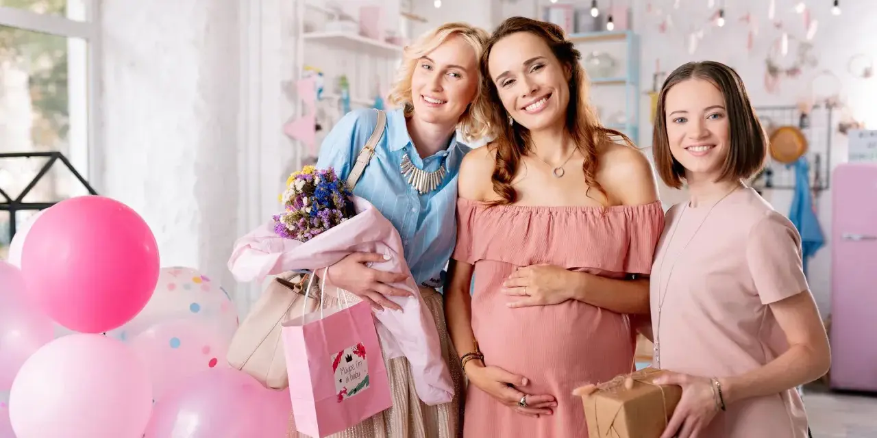 Trzy uśmiechnięte kobiety świętują baby shower. Jedna z nich jest w ciąży, a pozostałe dwie trzymają prezenty i balony.