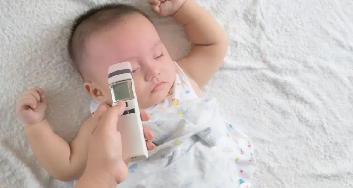 Ręka mierzy niemowlakowi temperaturę na czole termometrem bezdotykowym. Spokojny sen malucha to znak, że prawidłowa temperatura u niemowlaka na czole jest utrzymana.