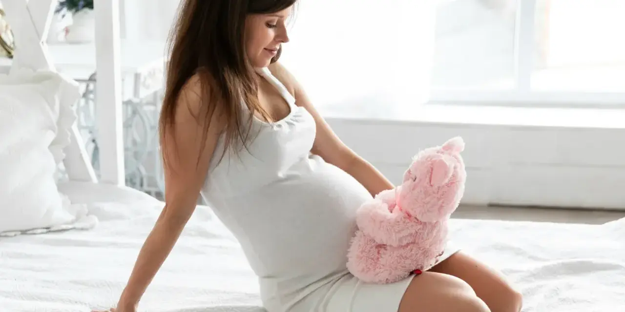 Szczęśliwa przyszła mama w 6 miesiącu ciąży, trzymająca różowego misia na brzuszku.