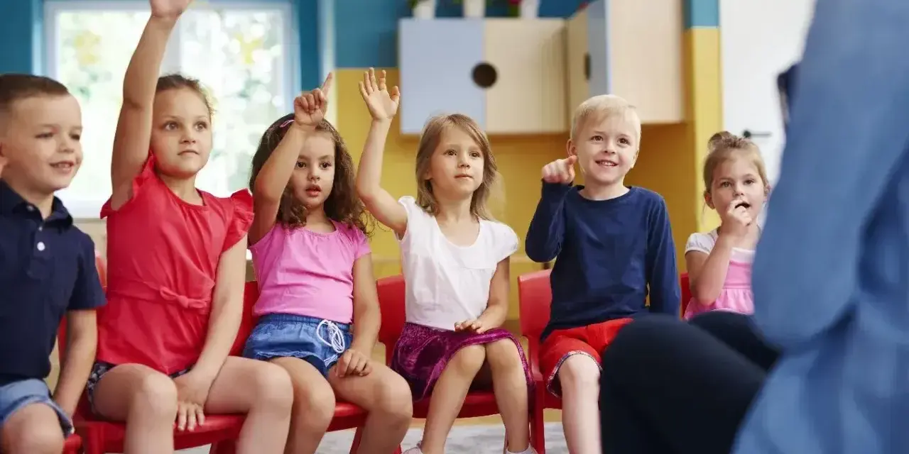 Dzieci na czerwonych krzesłach podnoszą ręce, jakby odpowiadały na pytanie, czy zerówka jest obowiązkowa.