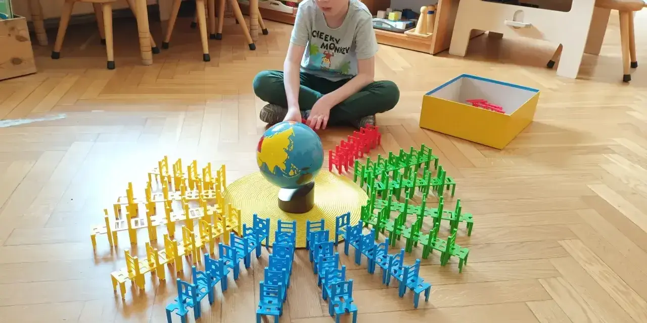 Chłopiec bawi się w przedszkolu Montessori, układając kolorowe krzesełka wokół globusa. To pokazuje, montessori co to za metoda nauczania.