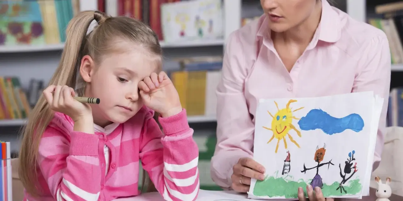 Dziewczynka z kredką w dłoni, smutna, podczas sesji socjoterapii. Terapeuta pokazuje jej rysunek.