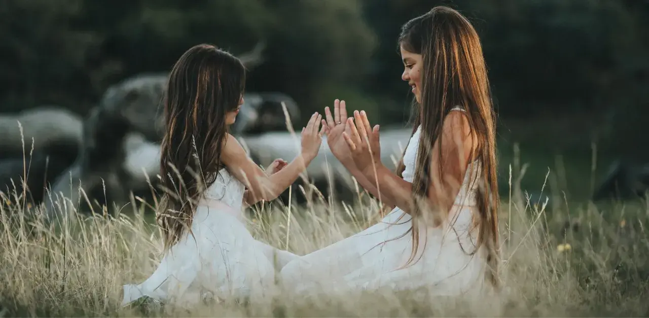 Dwie dziewczynki w białych sukienkach siedzą na łące i bawią się w "pajacyka". To idealna wyliczanka dla dzieci, która rozwija koordynację ruchową.