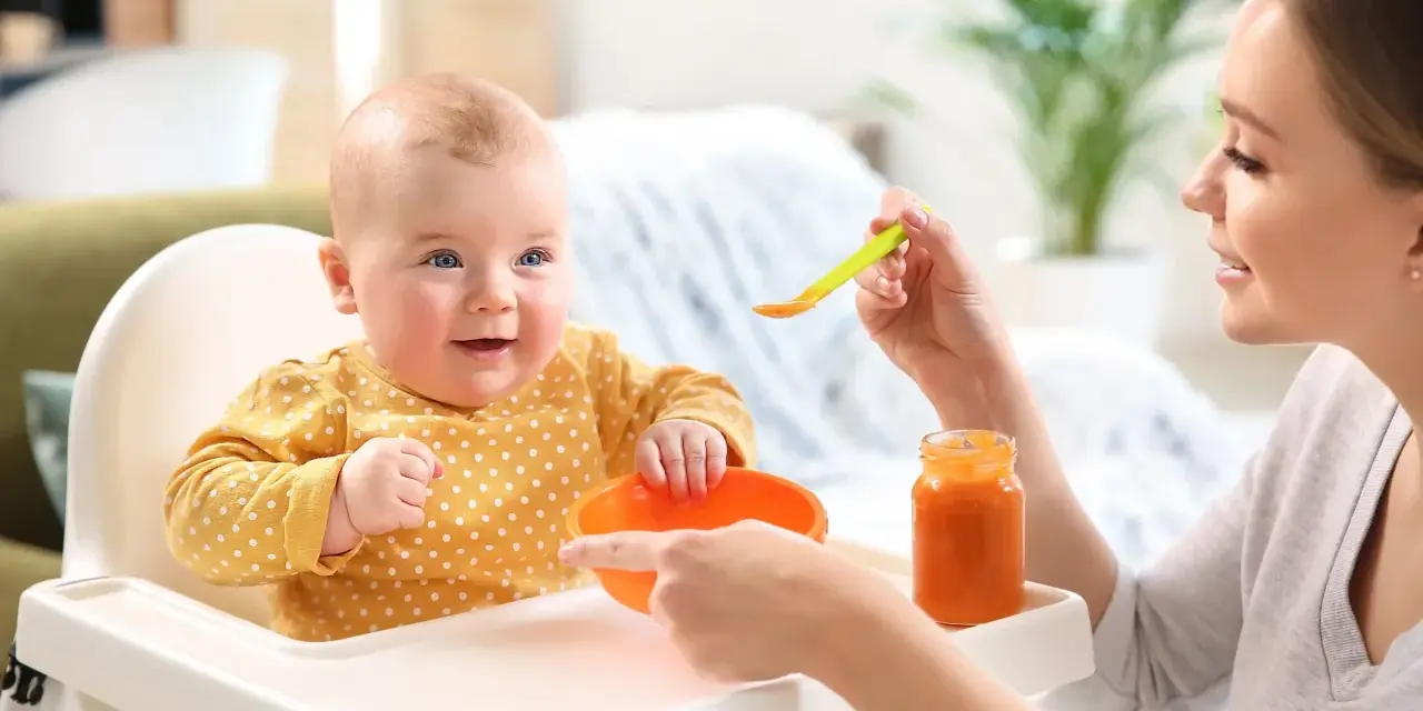 Uśmiechnięte 2 miesięczne dziecko w żółtym ubranku w kropki siedzi w krzesełku do karmienia. Mama podaje mu jedzenie z łyżeczki.