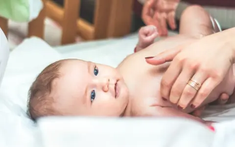 Niebieskooki bobas leży na białym prześcieradle, a jego mama delikatnie go dotyka. Na palcu mamy widać obrączkę, a na jej paznokciach ciemny lakier. Dziecko ma na sobie tylko pieluszkę, a jego skóra jest gładka i delikatna, jak **perły Epsteina**.