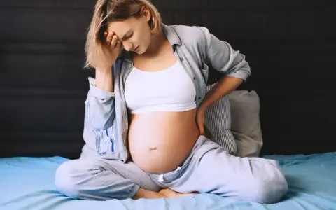 Ciężarna kobieta siedzi na łóżku, trzymając się za głowę i plecy. Odczuwa ból krocza w ciąży, co widać po jej wyrazie twarzy.