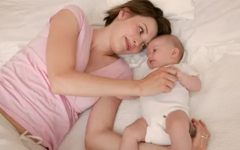 Mama patrzy z miłością na swoje 5 tygodniowe dziecko leżące obok niej na łóżku.
