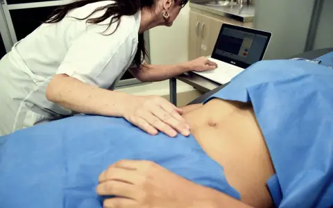 Lekarka bada brzuch pacjentki, patrząc na ekran laptopa z wykresami.