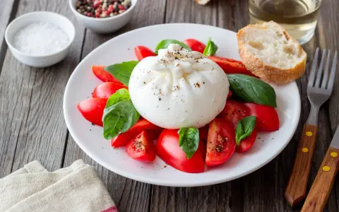 Burrata z pomidorami i bazylią na talerzu, obok pieczywo i przyprawy.