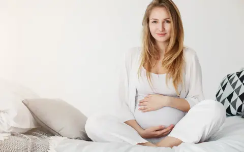 Uśmiechnięta kobieta w ciąży w białych ubraniach siedzi na łóżku, delikatnie trzymając się za brzuch.