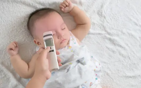 Ręka mierzy niemowlakowi temperaturę na czole termometrem bezdotykowym. Spokojny sen malucha to znak, że prawidłowa temperatura u niemowlaka na czole jest utrzymana.