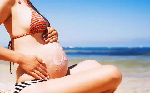 Ciężarna kobieta na plaży, z kremem przeciwsłonecznym na brzuchu w kształcie słońca. Bezpieczne opalanie natryskowe w ciąży.