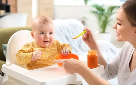 Uśmiechnięte 2 miesięczne dziecko w żółtym ubranku w kropki siedzi w krzesełku do karmienia. Mama podaje mu jedzenie z łyżeczki.