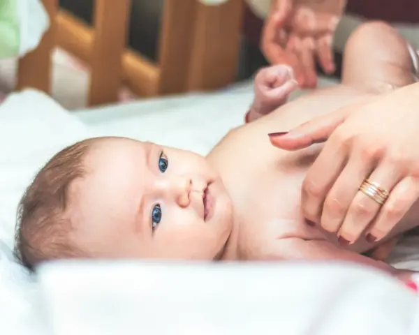 Niebieskooki bobas leży na białym prześcieradle, a jego mama delikatnie go dotyka. Na palcu mamy widać obrączkę, a na jej paznokciach ciemny lakier. Dziecko ma na sobie tylko pieluszkę, a jego skóra jest gładka i delikatna, jak **perły Epsteina**.