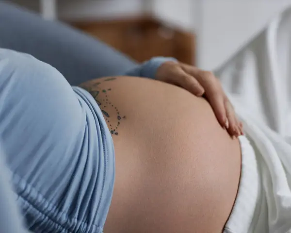 Dłoń delikatnie spoczywa na **twardym brzuchu w ciąży**, ozdobionym subtelnym tatuażem.