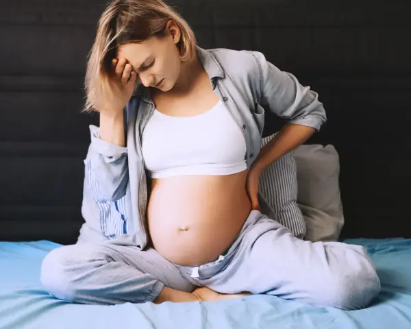 Ciężarna kobieta siedzi na łóżku, trzymając się za głowę i plecy. Odczuwa ból krocza w ciąży, co widać po jej wyrazie twarzy.