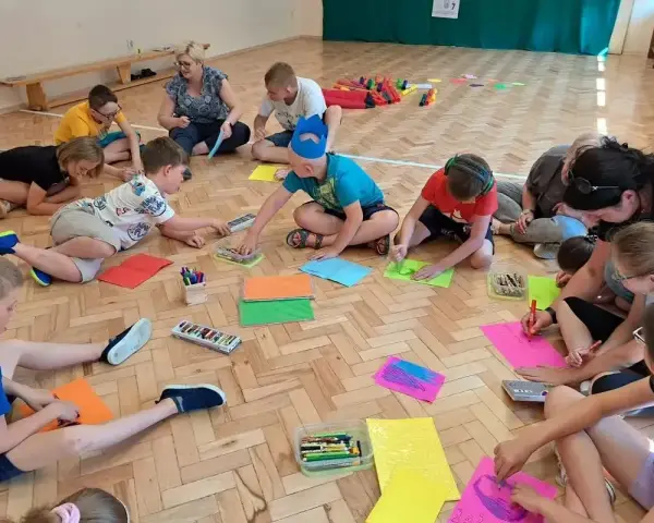 Dzieci i dorośli siedzą na podłodze, rysując i bawiąc się. Wszyscy są zaangażowani w zabawy integracyjne, tworząc kolorowe prace.
