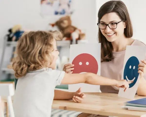 Dziecko i pedagog pracują nad emocjami podczas terapii pedagogicznej, używając kart z buźkami.