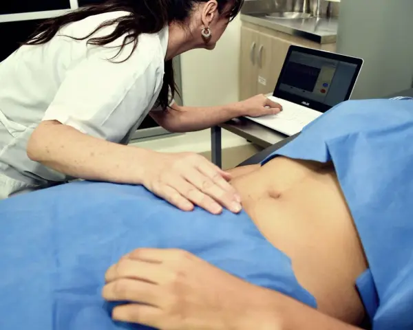 Lekarka bada brzuch pacjentki, patrząc na ekran laptopa z wykresami.