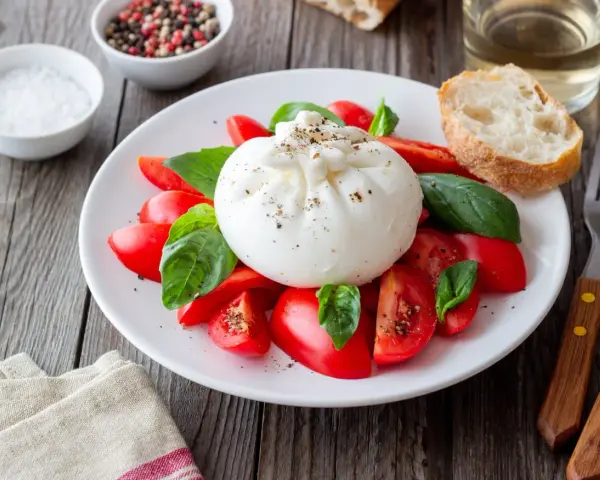Burrata z pomidorami i bazylią na talerzu, obok pieczywo i przyprawy.