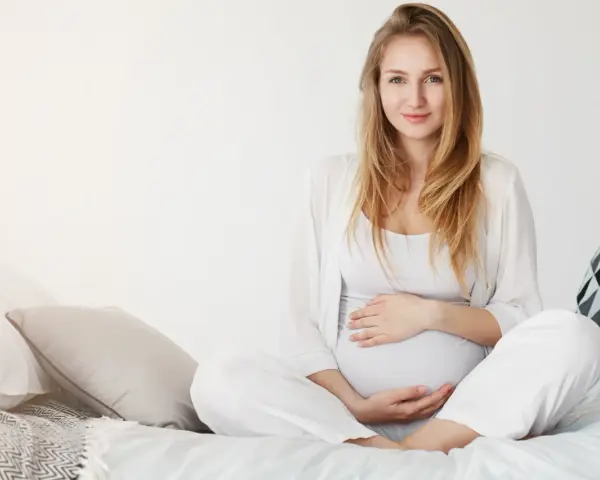 Uśmiechnięta kobieta w ciąży w białych ubraniach siedzi na łóżku, delikatnie trzymając się za brzuch.