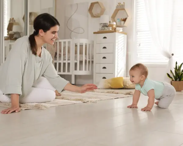 Uśmiechnięta mama zachęca do pierwszych kroków, kiedy dziecko raczkuje po dywanie w pokoju dziecięcym.