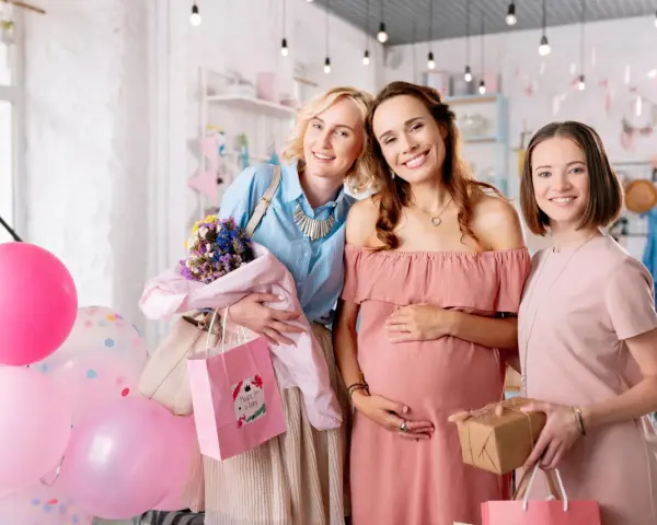 Trzy uśmiechnięte kobiety świętują baby shower. Jedna z nich jest w ciąży, a pozostałe dwie trzymają prezenty i balony.