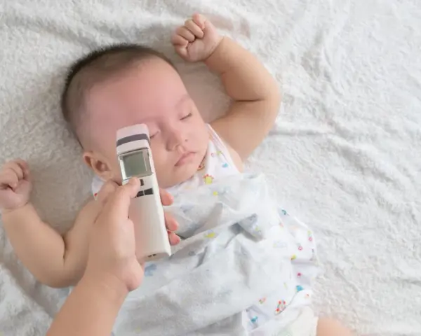 Ręka mierzy niemowlakowi temperaturę na czole termometrem bezdotykowym. Spokojny sen malucha to znak, że prawidłowa temperatura u niemowlaka na czole jest utrzymana.