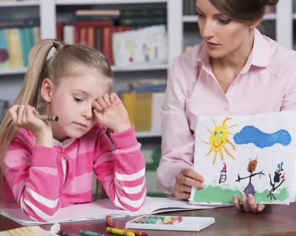 Dziewczynka z kredką w dłoni, smutna, podczas sesji socjoterapii. Terapeuta pokazuje jej rysunek.