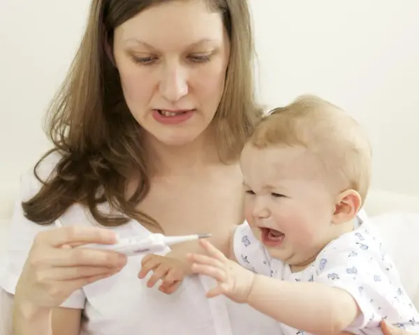 Zmartwiona mama mierzy temperaturę płaczącemu dziecku, które ma gorączkę po szczepieniu.