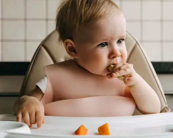 Małe dziecko w różowym śliniaku próbuje kawałek marchewki. Na tacce leżą kolejne pomarańczowe kawałki.