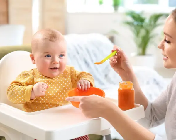 Uśmiechnięte 2 miesięczne dziecko w żółtym ubranku w kropki siedzi w krzesełku do karmienia. Mama podaje mu jedzenie z łyżeczki.