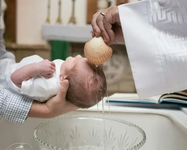 Chrzestna polewa główkę dziecka wodą święconą z muszli podczas ceremonii chrztu.