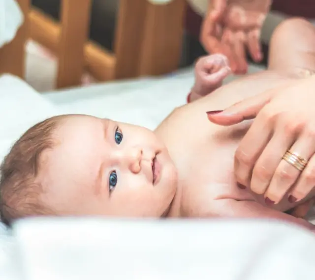 Niebieskooki bobas leży na białym prześcieradle, a jego mama delikatnie go dotyka. Na palcu mamy widać obrączkę, a na jej paznokciach ciemny lakier. Dziecko ma na sobie tylko pieluszkę, a jego skóra jest gładka i delikatna, jak **perły Epsteina**.